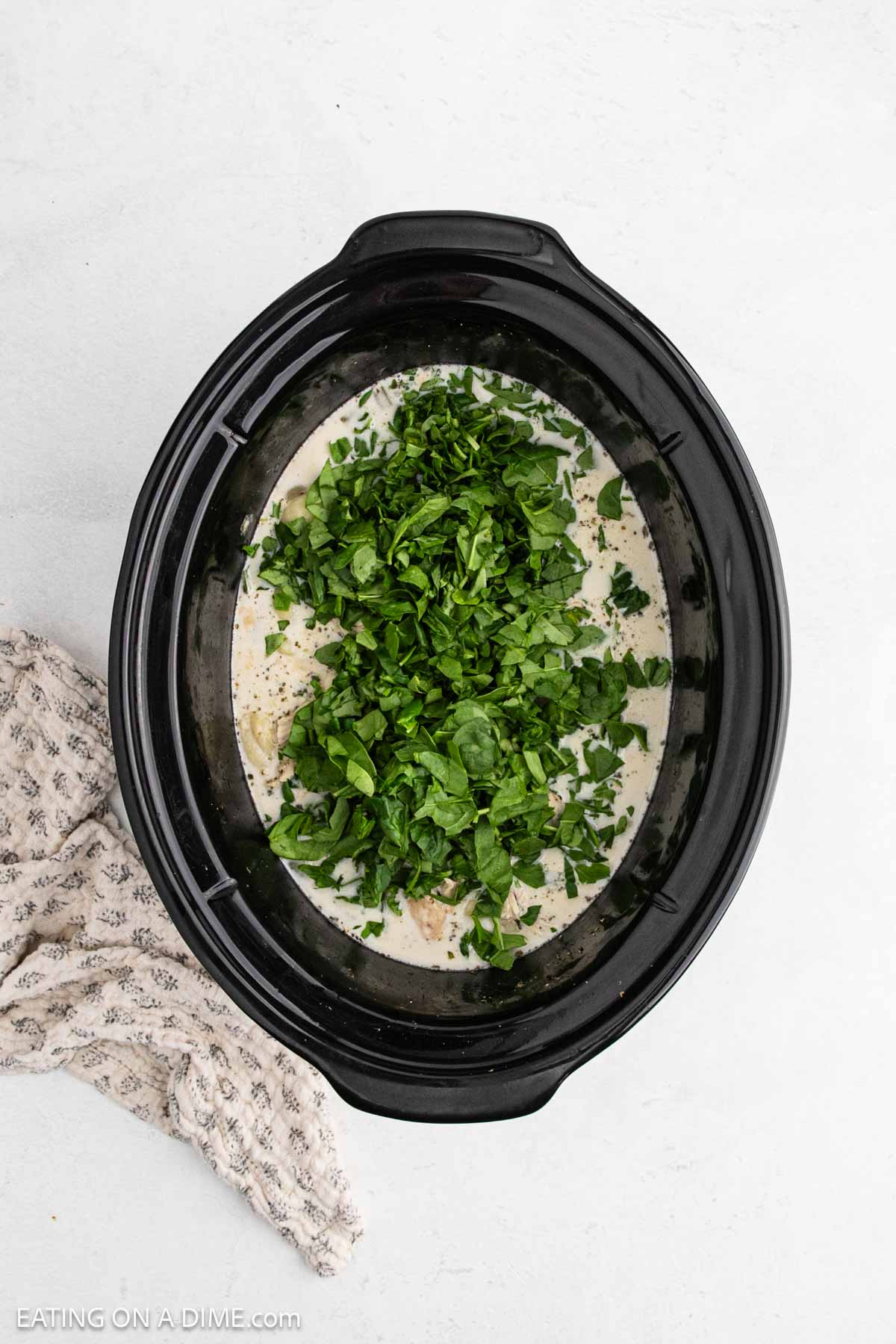A black Crock Pot filled with creamy Chicken Gnocchi Soup, topped with freshly chopped spinach, sits on a light surface next to a textured beige cloth.