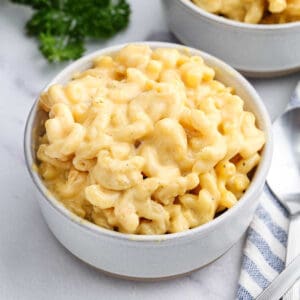 A white bowl filled with creamy Velveeta Mac and Cheese sits on a marble surface, with another bowl in the background and a striped napkin nearby.