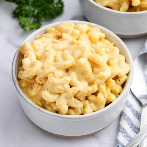 A white bowl filled with creamy Velveeta Mac and Cheese sits on a marble surface, with another bowl in the background and a striped napkin nearby.