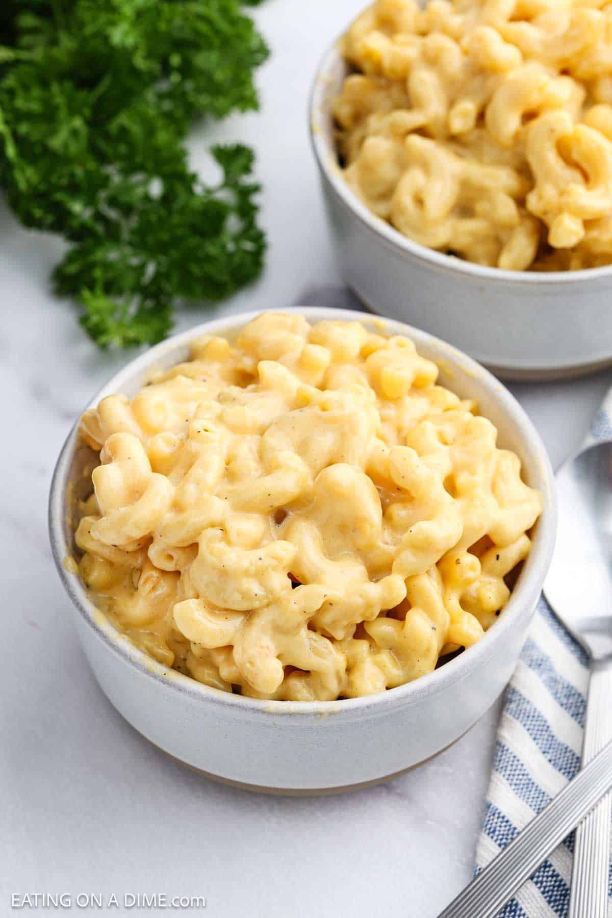 A bowl of creamy Velveeta Mac and Cheese sits on a white surface, with a second bowl partially visible in the background and fresh parsley nearby.