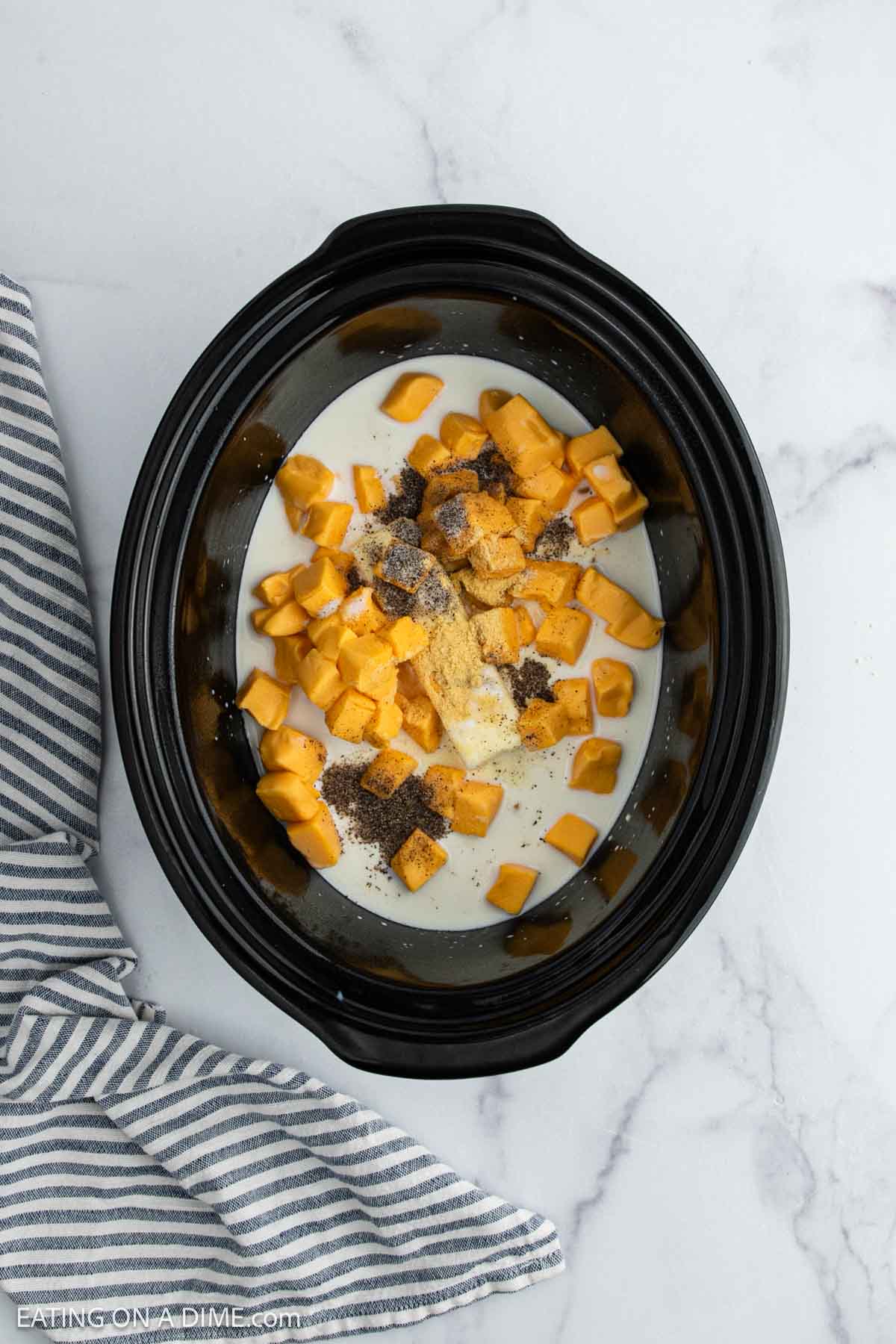 A Crock Pot filled with cubed potatoes, milk, seasonings, and spices sits on a marble countertop next to a gray and white striped cloth.