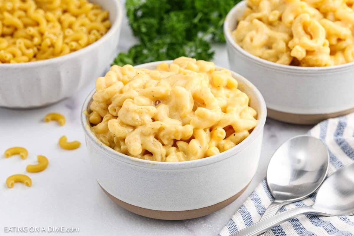 A bowl of creamy Velveeta Mac and Cheese sits on a white surface, with two more bowls in the background. A few uncooked macaroni pieces, two spoons, and a blue-striped napkin are nearby. Green herbs are visible in the background.