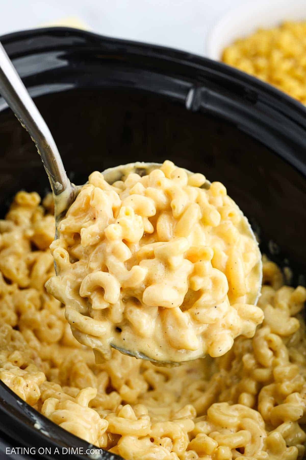 A ladle scoops creamy Mac and Cheese from a black Crock Pot, revealing cheesy, Velveeta-filled pasta in a rich, saucy layer.