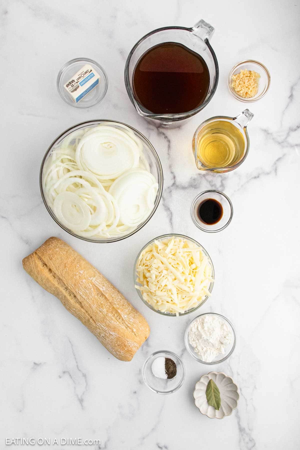 Overhead view of classic French Onion Soup ingredients on a white marble surface: sliced onions, beef broth, minced garlic, butter, olive oil, Worcestershire sauce, shredded cheese, flour, black pepper, bay leaves, and a baguette.