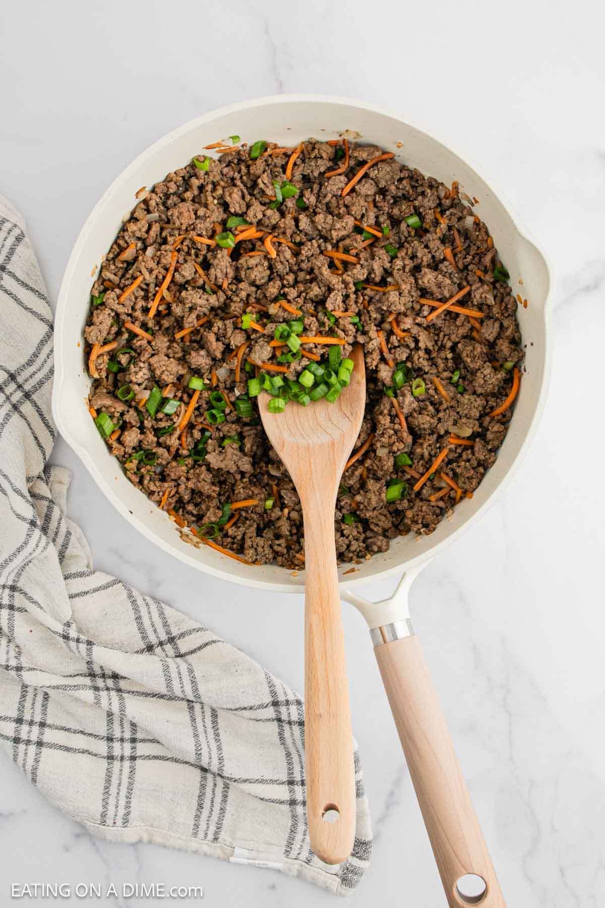 Cooked ground beef in a skillet with shredded carrots and chopped green onions with a wooden spoon