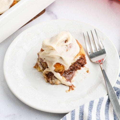 A single Hawaiian Roll Cinnamon Roll topped with creamy white icing sits on a white plate with a fork beside it, next to a striped cloth napkin.