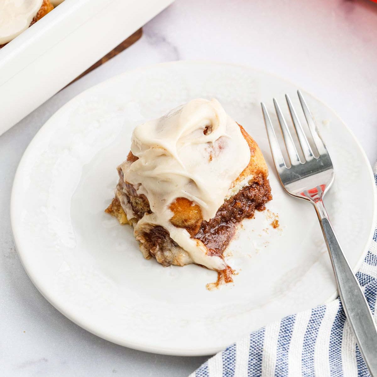 A single Hawaiian Roll Cinnamon Roll topped with creamy white icing sits on a white plate with a fork beside it, next to a striped cloth napkin.