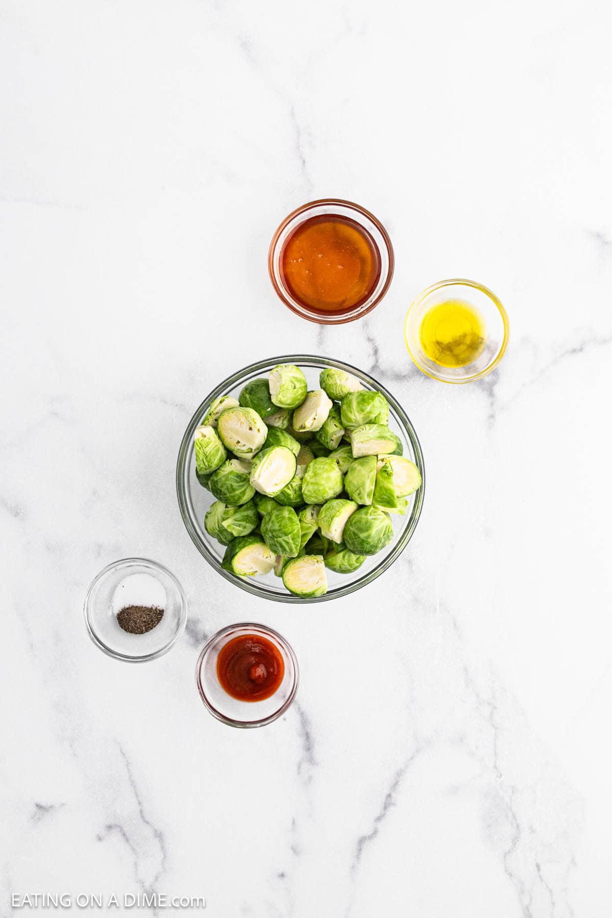 A bowl of halved Brussels sprouts is surrounded by small bowls of olive oil, honey, hot sauce, and ground pepperโperfect for making zesty Honey Sriracha Brussel Sprouts on a white marble surface.