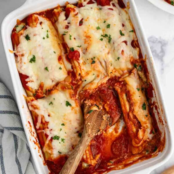 A baking dish filled with cheesy baked lasagna and manicotti, with a wooden spoon resting inside and one piece partially served. The lasagna is topped with melted cheese and garnished with chopped herbs.