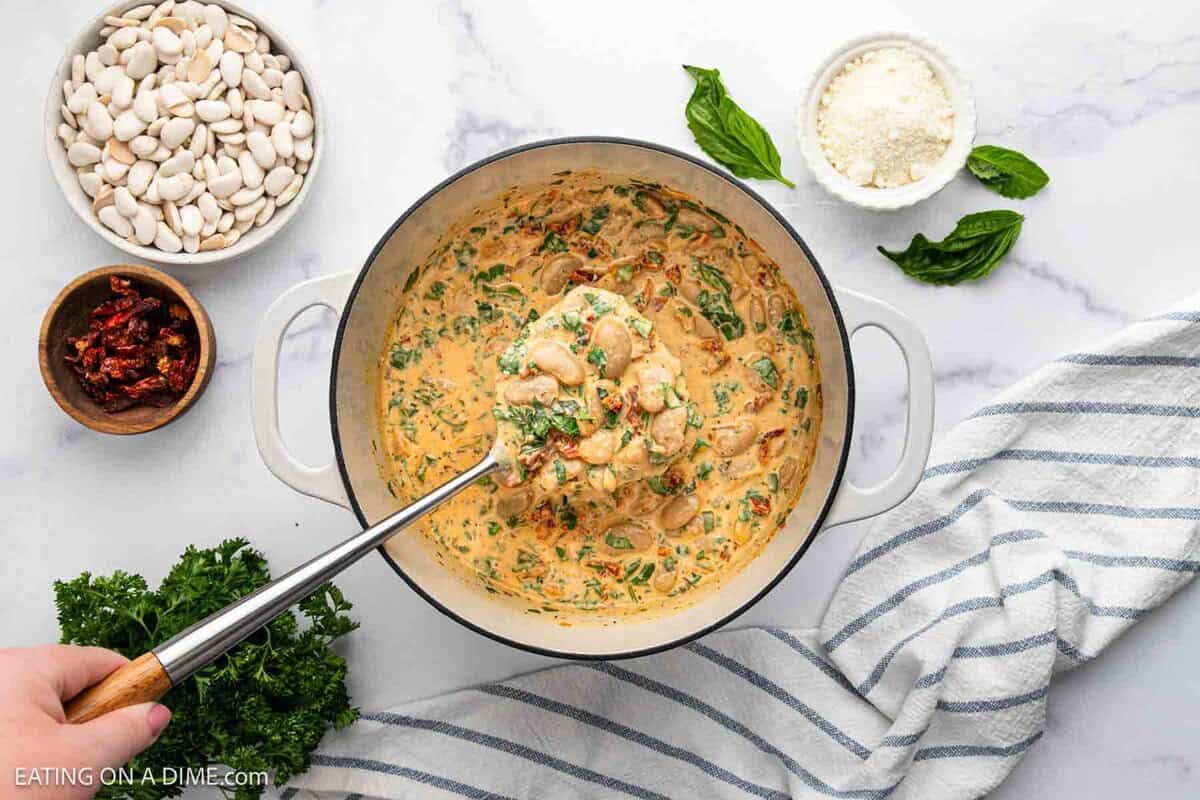 Overhead view of a creamy Marry Me Butter Beans and spinach dish in a pot with a ladle, surrounded by bowls of white beans, grated cheese, sun-dried tomatoes, and fresh basil on a marble countertop with a striped towel.