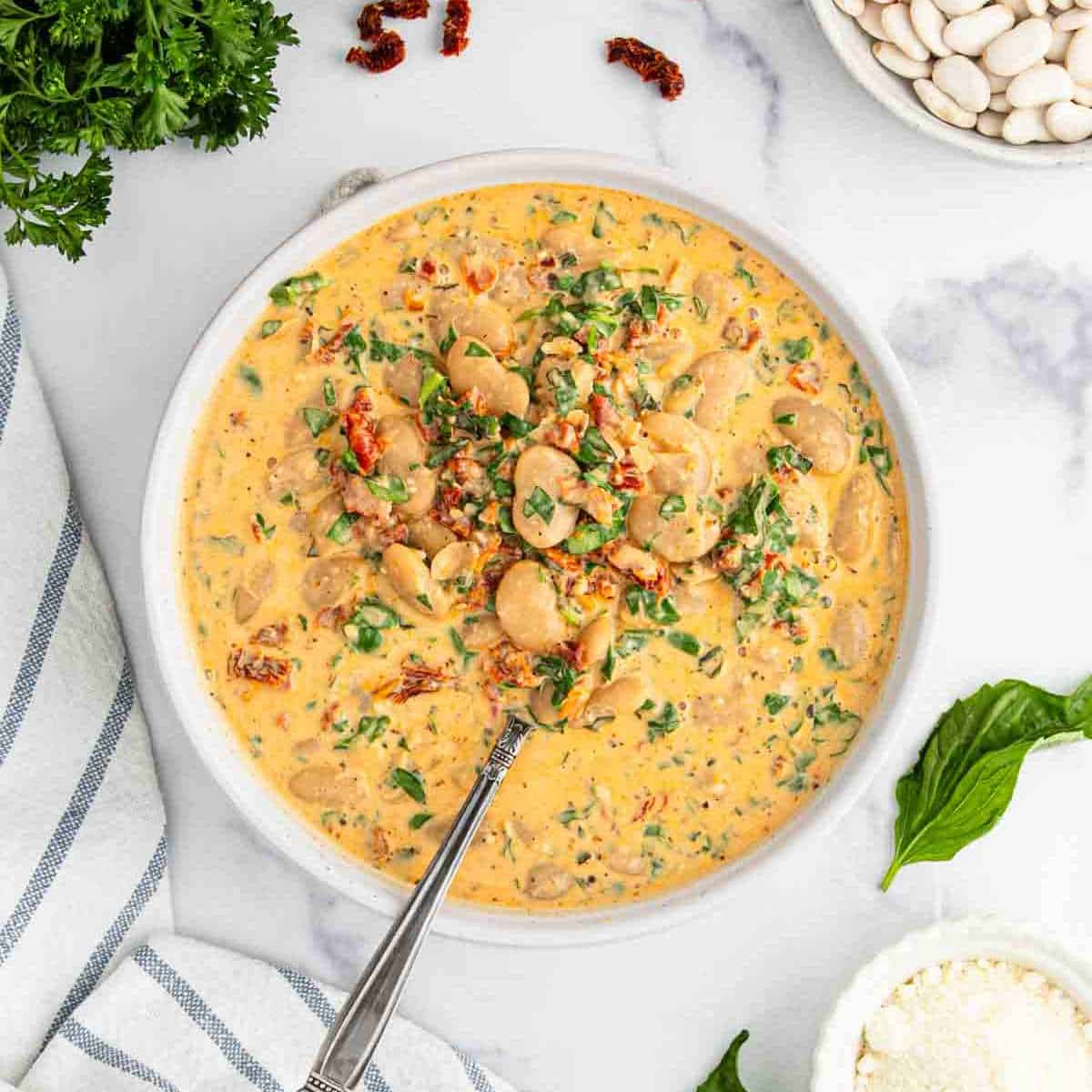 A bowl of creamy Marry Me Butter Beans stew garnished with chopped herbs and sun-dried tomatoes, with a spoon in the bowl. Fresh parsley, basil, white beans, and grated cheese are artfully arranged on a marble surface.