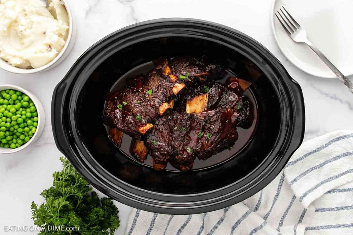A black slow cooker filled with tender Slow Cooker Beef Ribs in sauce, garnished with herbs. Nearby are bowls of peas and mashed potatoes, a fork, and a bunch of fresh parsley on a white surface.
