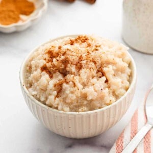 A white bowl filled with creamy Slow Cooker Rice Pudding, topped with a sprinkle of ground cinnamon, sits on a marble surface next to a spoon and a striped napkin.