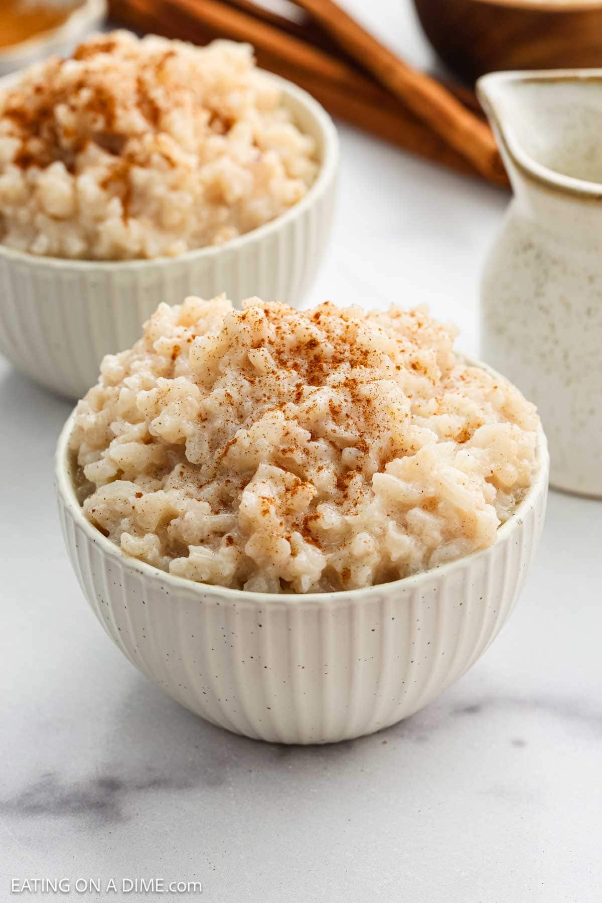 A white bowl filled with creamy Slow Cooker Rice Pudding, sprinkled with cinnamon on top, sits on a white surface. Another bowl of rice pudding and a small pitcher are blurred in the background.