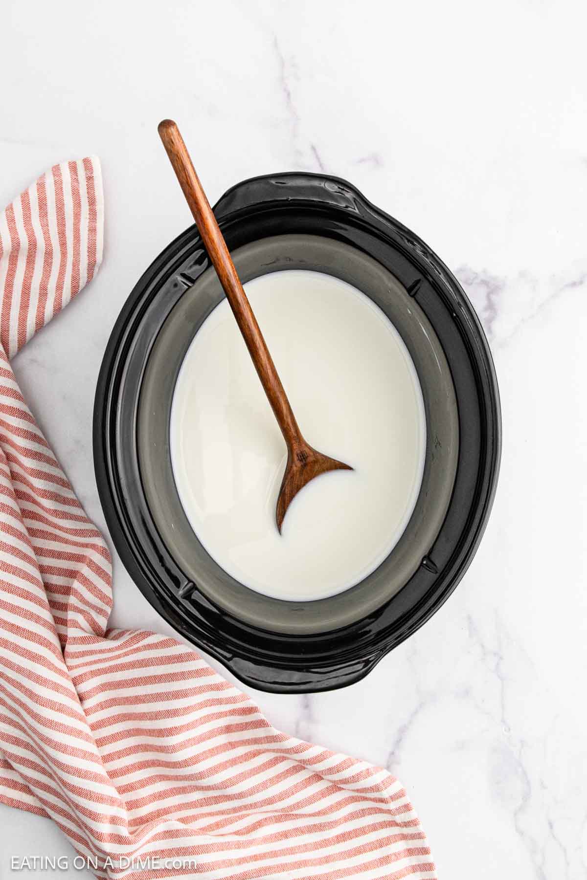 A slow cooker filled with creamy rice pudding and stirred by a wooden spoon sits on a white marble surface next to a red and white striped cloth.