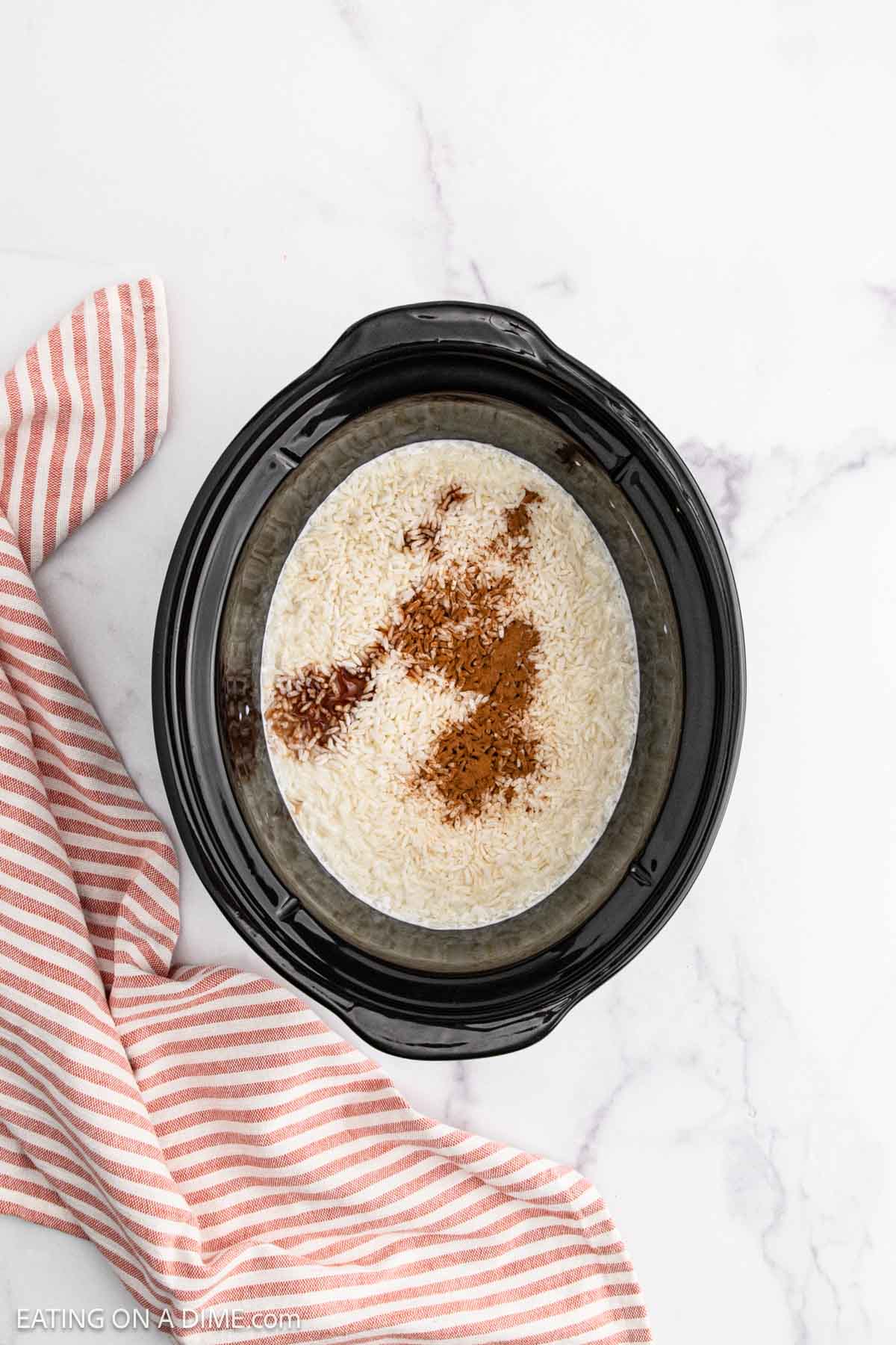 A slow cooker with rice, cinnamon, and liquid inside, perfect for making Slow Cooker Rice Pudding, sits on a white marble surface next to a red and white striped kitchen towel.