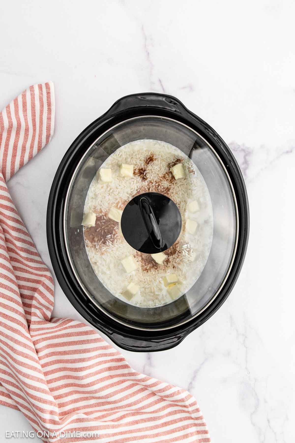 A slow cooker ready for Slow Cooker Rice Pudding is filled with uncooked rice, small cubes of butter, and seasonings, sitting on a white marble surface next to a red and white striped kitchen towel.