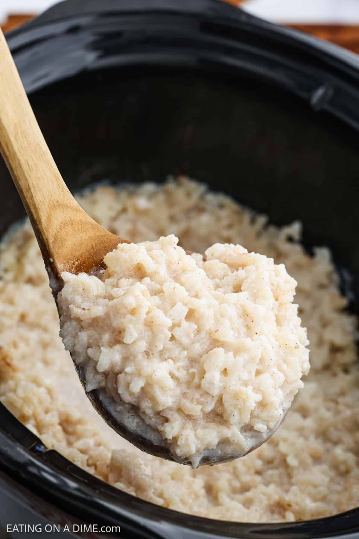 A wooden spoon holds a serving of creamy Slow Cooker Rice Pudding above a slow cooker filled with more of the thick, textured dessert.