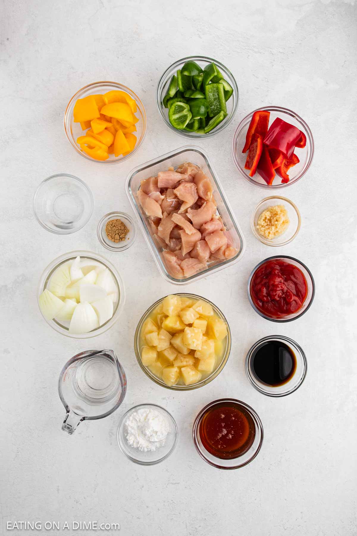 A top-down view of bowls with diced bell peppers, cubed raw chicken, minced garlic, ketchup, soy sauce, pineapple chunks, chopped onion, brown sugar, cornstarch, water, and sweet and sour sauce—perfect for a Sweet and Sour Chicken recipe.