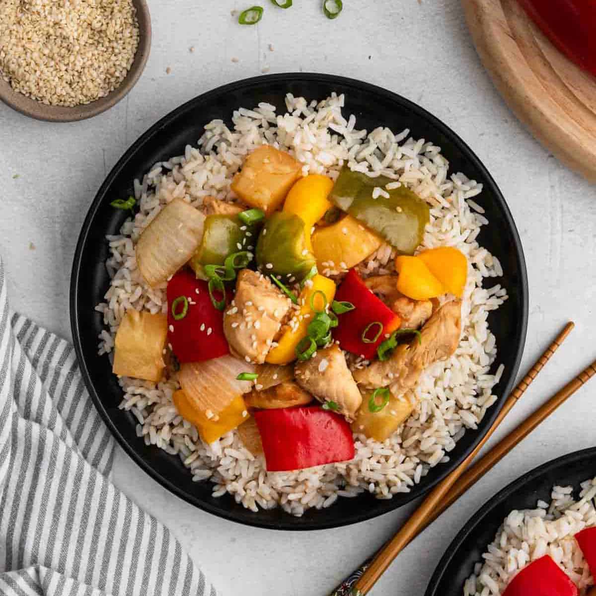 A black plate with white rice topped with sweet and sour chicken recipe, featuring pieces of chicken, red and green bell peppers, pineapple, and onion, garnished with sesame seeds and chopped green onions. Chopsticks and a striped napkin are beside the plate.