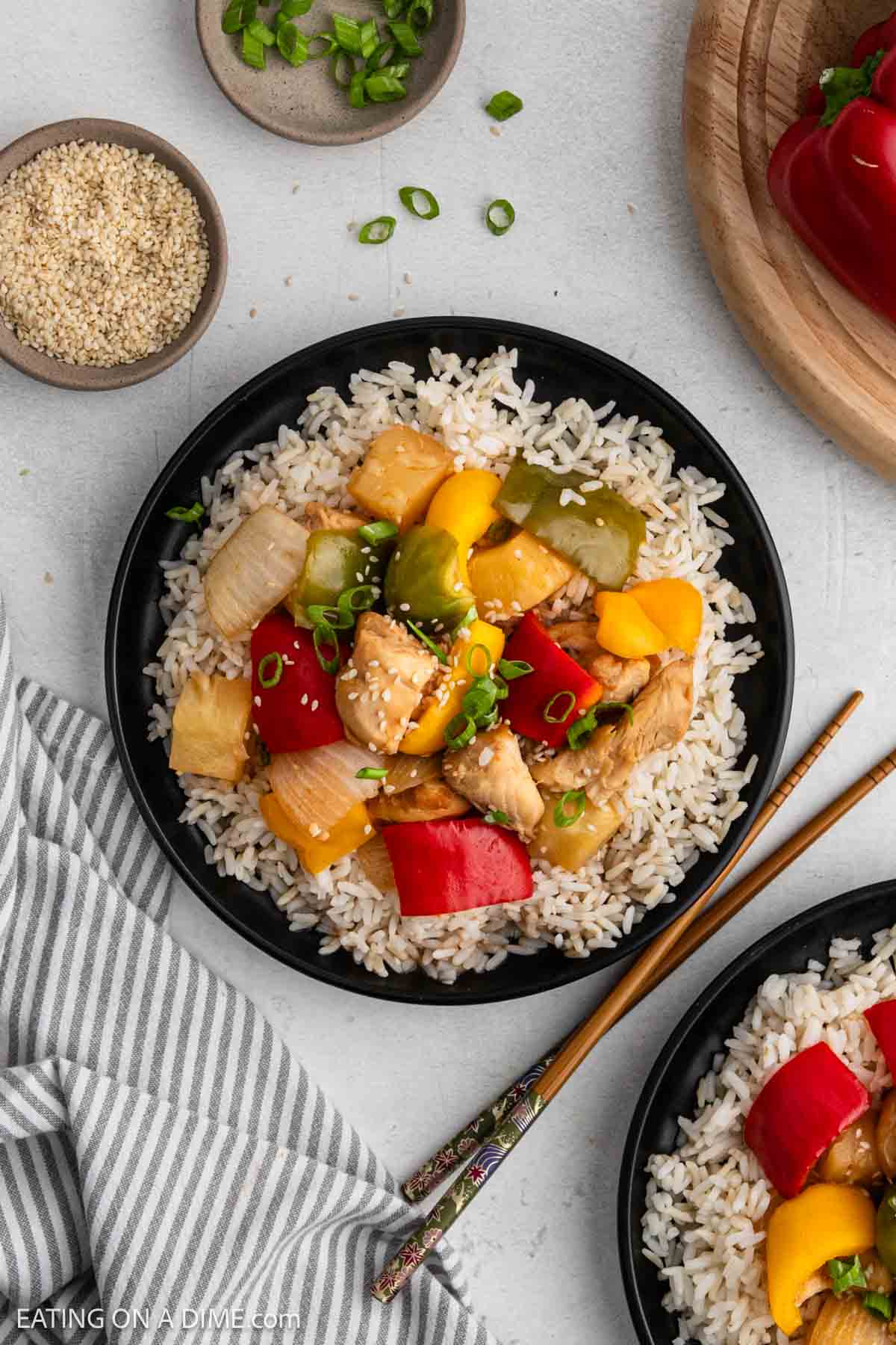 A black plate filled with white rice topped with colorful slow cooker sweet and sour chicken, pineapple, bell peppers, onions, and garnished with sesame seeds and chopped green onions. Chopsticks rest beside the plate.