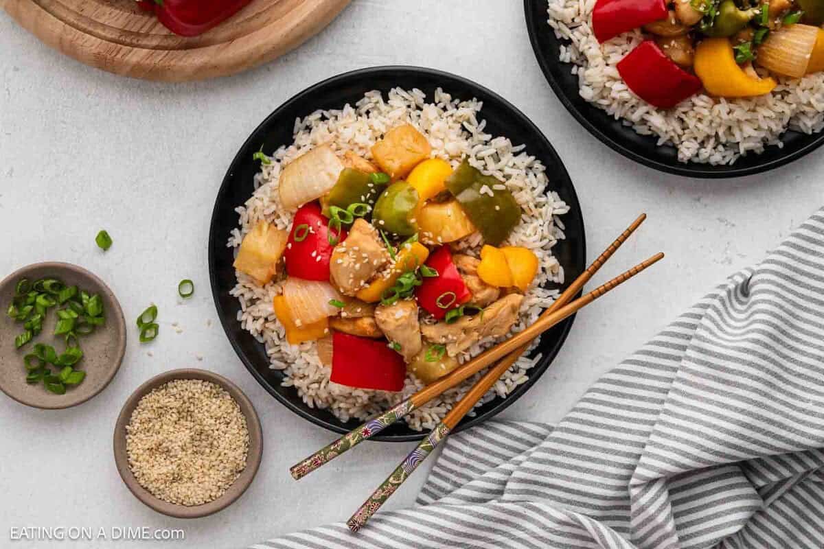 A plate of brown rice topped with chunks of Slow Cooker Sweet and Sour Chicken, red and yellow bell peppers, green peppers, and onions. Chopsticks rest on the plate, with small bowls of sliced green onions and sesame seeds nearby.