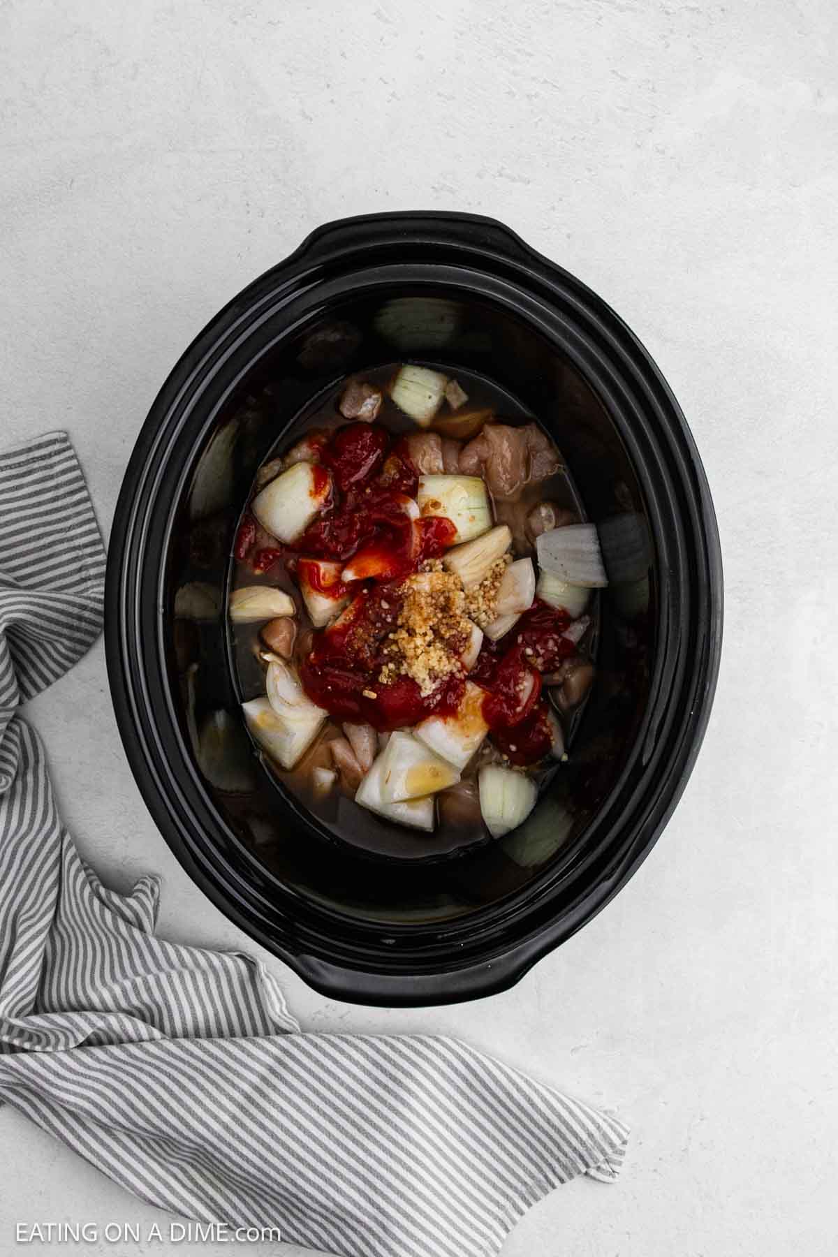 Overhead view of a slow cooker filled with chopped onions, raw chicken pieces for sweet and sour chicken, diced tomatoes, and minced garlic, placed on a light surface next to a gray striped cloth.