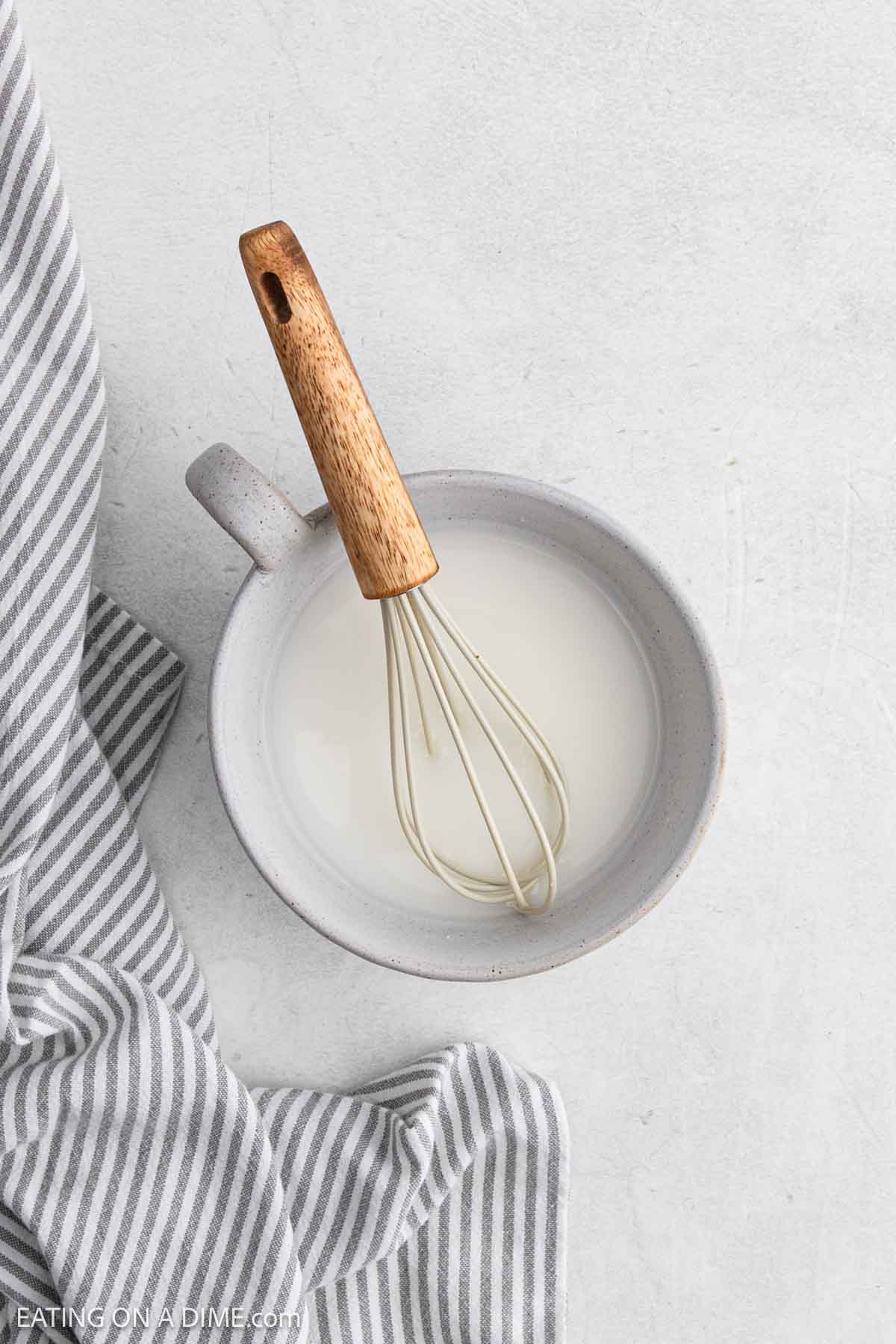 A ceramic bowl filled with milk and a whisk with a wooden handle sits on a white surface next to a gray and white striped cloth, ready for mixing ingredients for your favorite Sweet and Sour Chicken recipe.