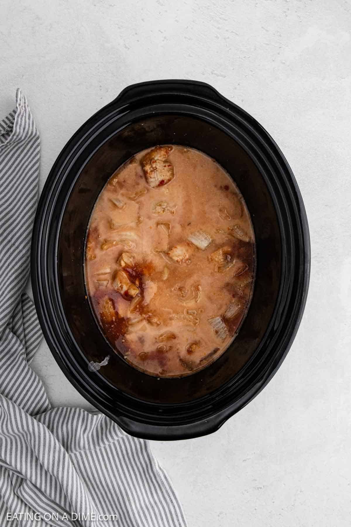 A black slow cooker filled with pieces of meat in a creamy, light brown sauce sits on a light surface next to a gray and white striped kitchen towel—perfect for trying your next chicken recipe.