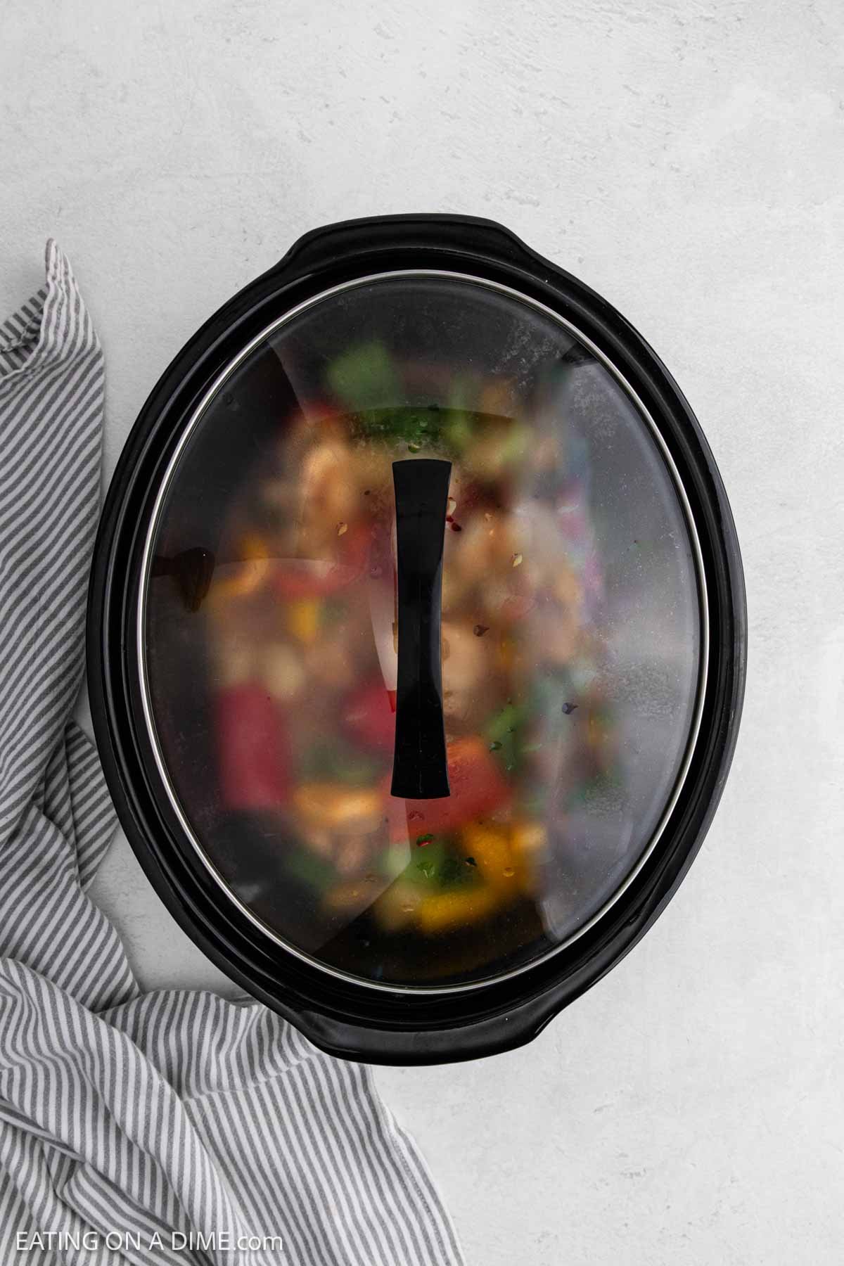 A black slow cooker with a glass lid, partially fogged up, reveals colorful Sweet and Sour Chicken inside. A gray and white striped kitchen towel rests next to it on a light gray surface.