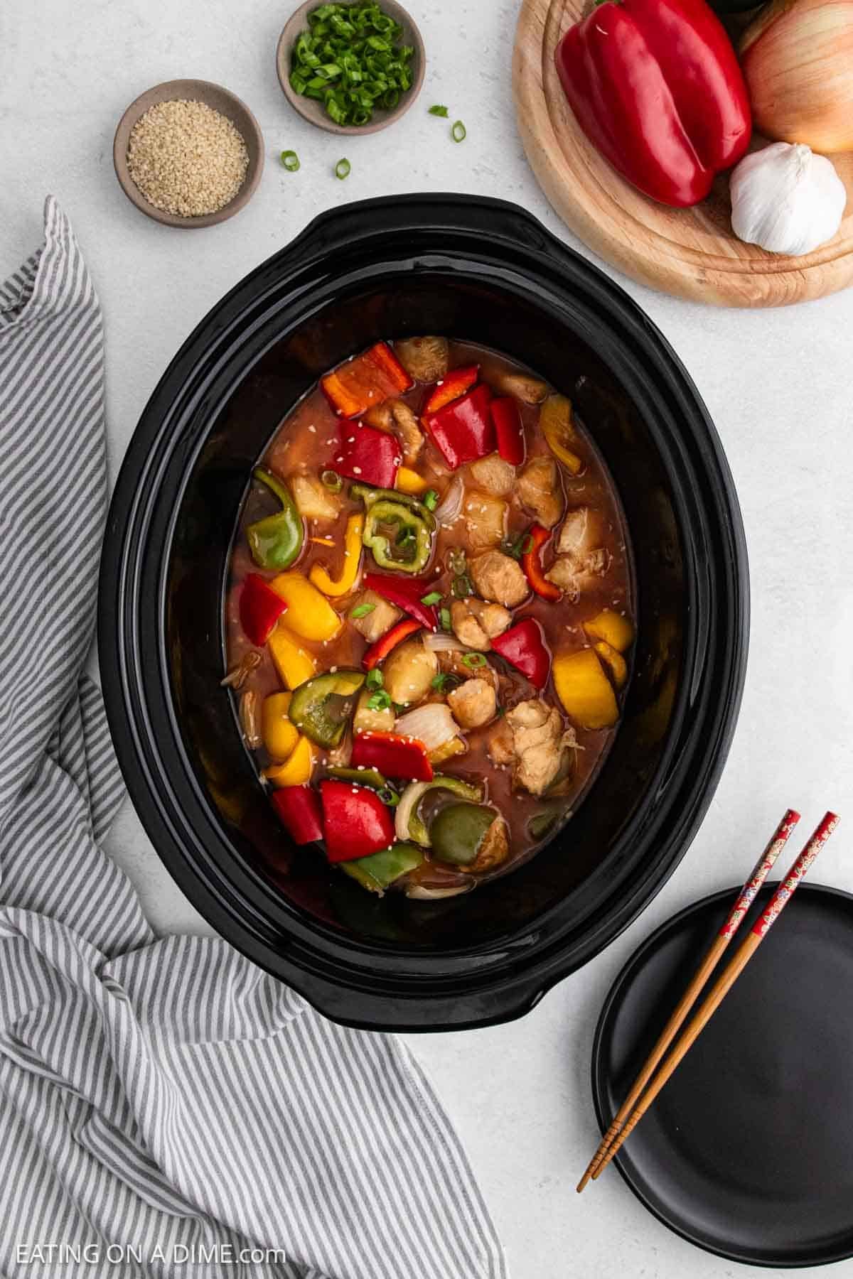 A slow cooker filled with Sweet and Sour Chicken, bell peppers, and sauce sits on a white surface. Surrounding it are a striped napkin, black plate with chopsticks, and bowls of sesame seeds and green onions.