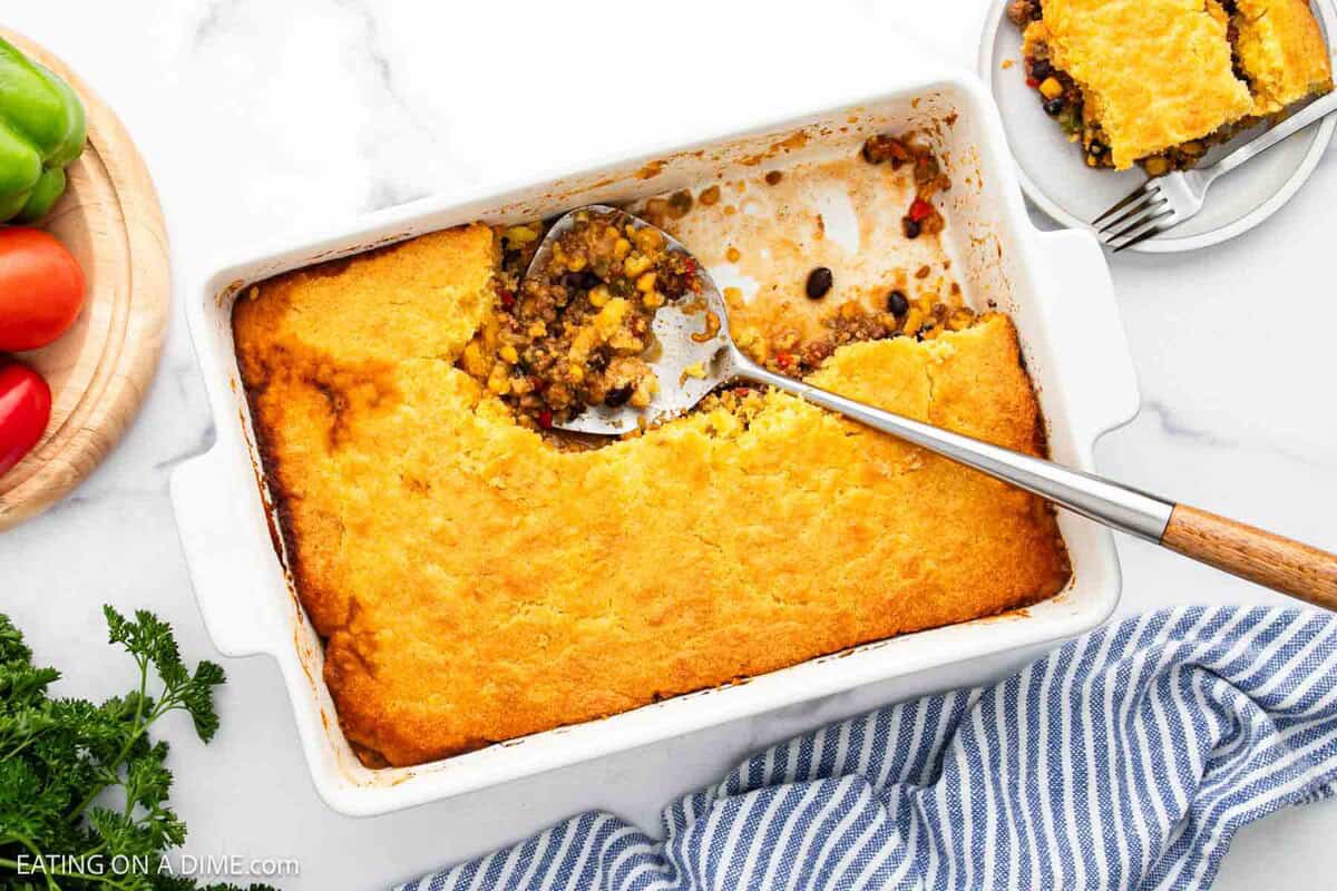 A casserole dish with a golden cornbread topping sits on a white surface. A serving has been scooped out, revealing a filling of beans, corn, and vegetables. A spoon rests inside, and a plate with a portion is nearby.