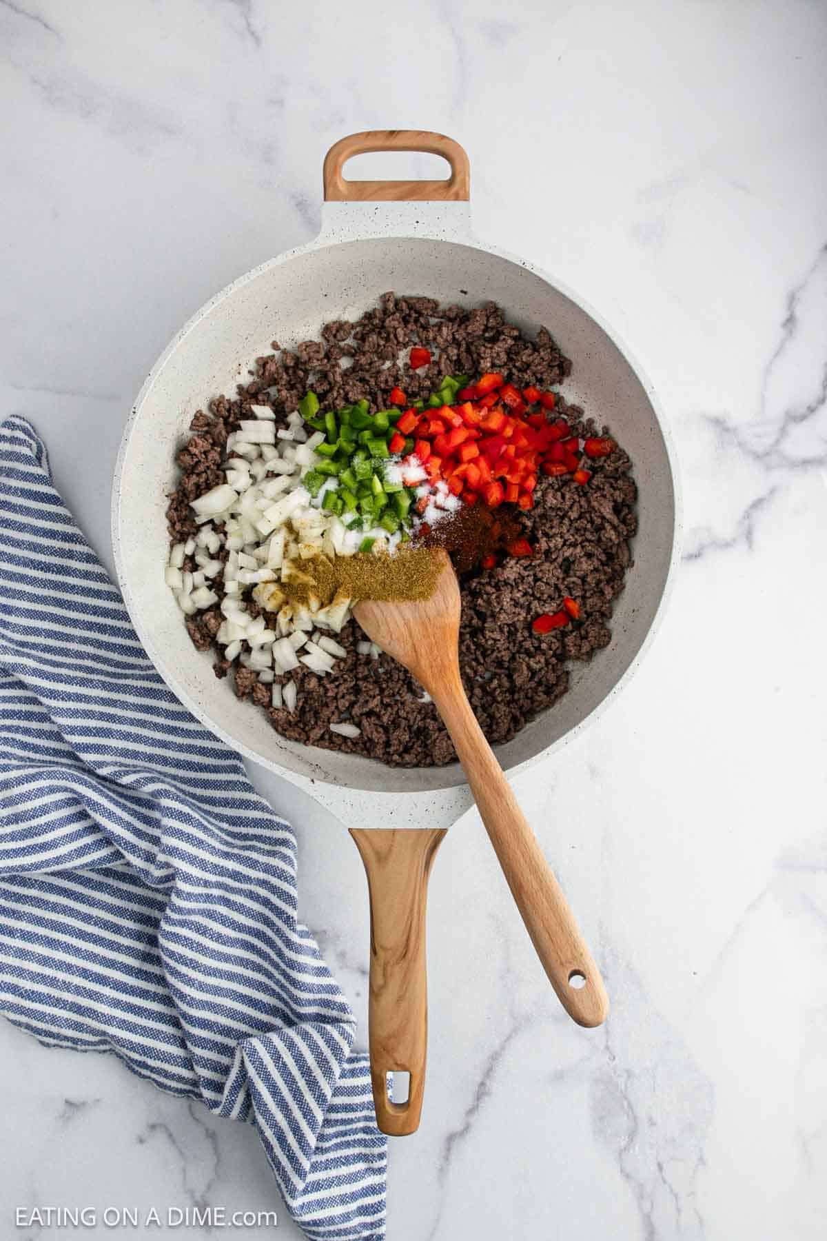 A skillet with cooked ground beef, topped with chopped onions, green and red bell peppers, and seasonings sits on a marble countertop next to a striped blue and white towel. A wooden spoon rests in the skillet.