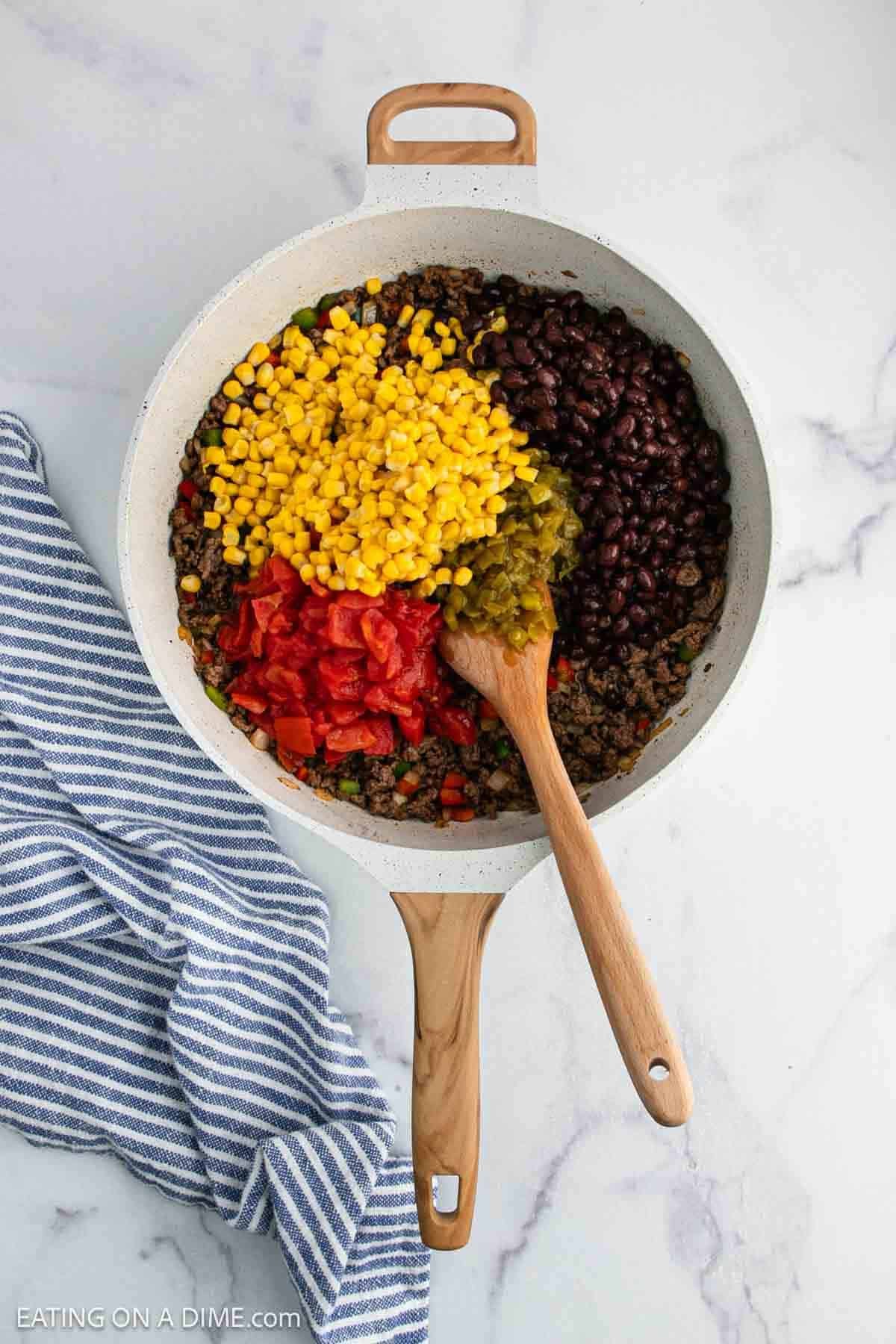 A skillet filled with ground beef, black beans, corn, diced tomatoes, and green chiles being mixed with a wooden spoon. A blue and white striped kitchen towel is beside the skillet on a marble countertop.