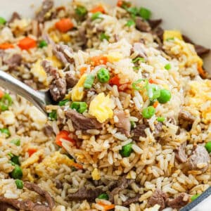 A close-up of a spoonful of Beef Fried Rice with beef strips, scrambled eggs, peas, carrots, and green onions, held above a bowl filled with the same colorful mixture.