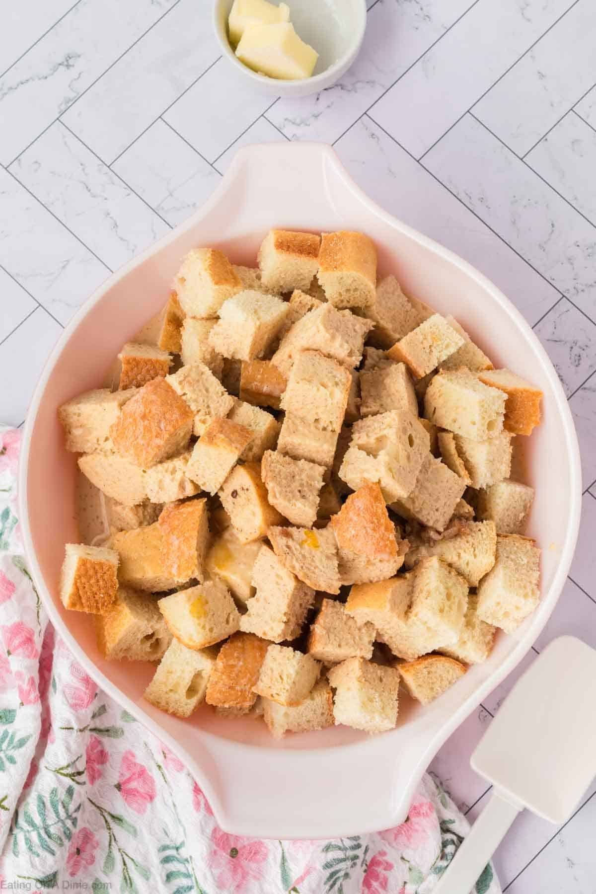 A pink bowl filled with cubed pieces of bread for bread pudding sits on a white tiled surface, next to a floral-patterned cloth, a white spatula, and a small bowl of butter.