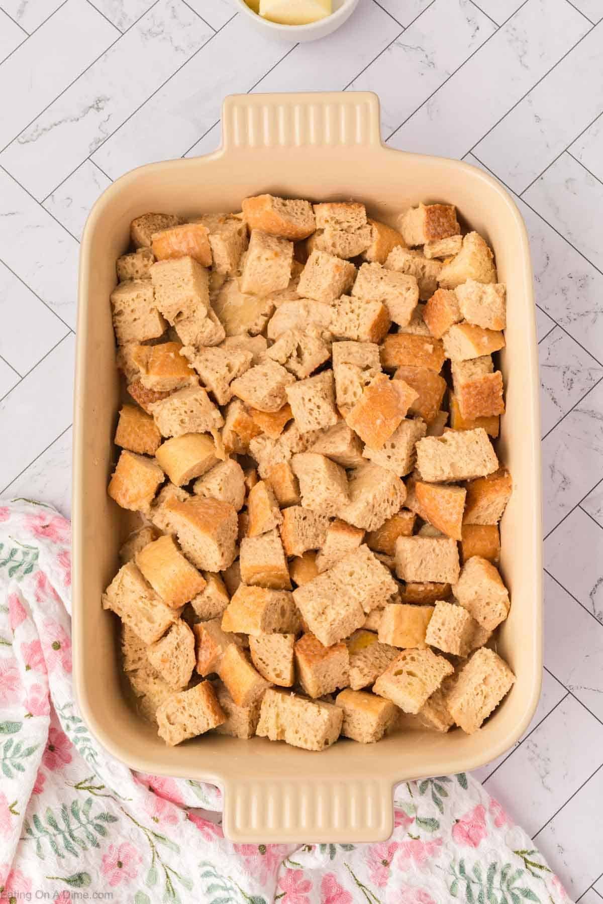 A beige baking dish filled with cubed bread pieces, ready to become a delicious bread pudding, sits on a white tiled surface, with a pink floral cloth partially visible in the bottom left corner.