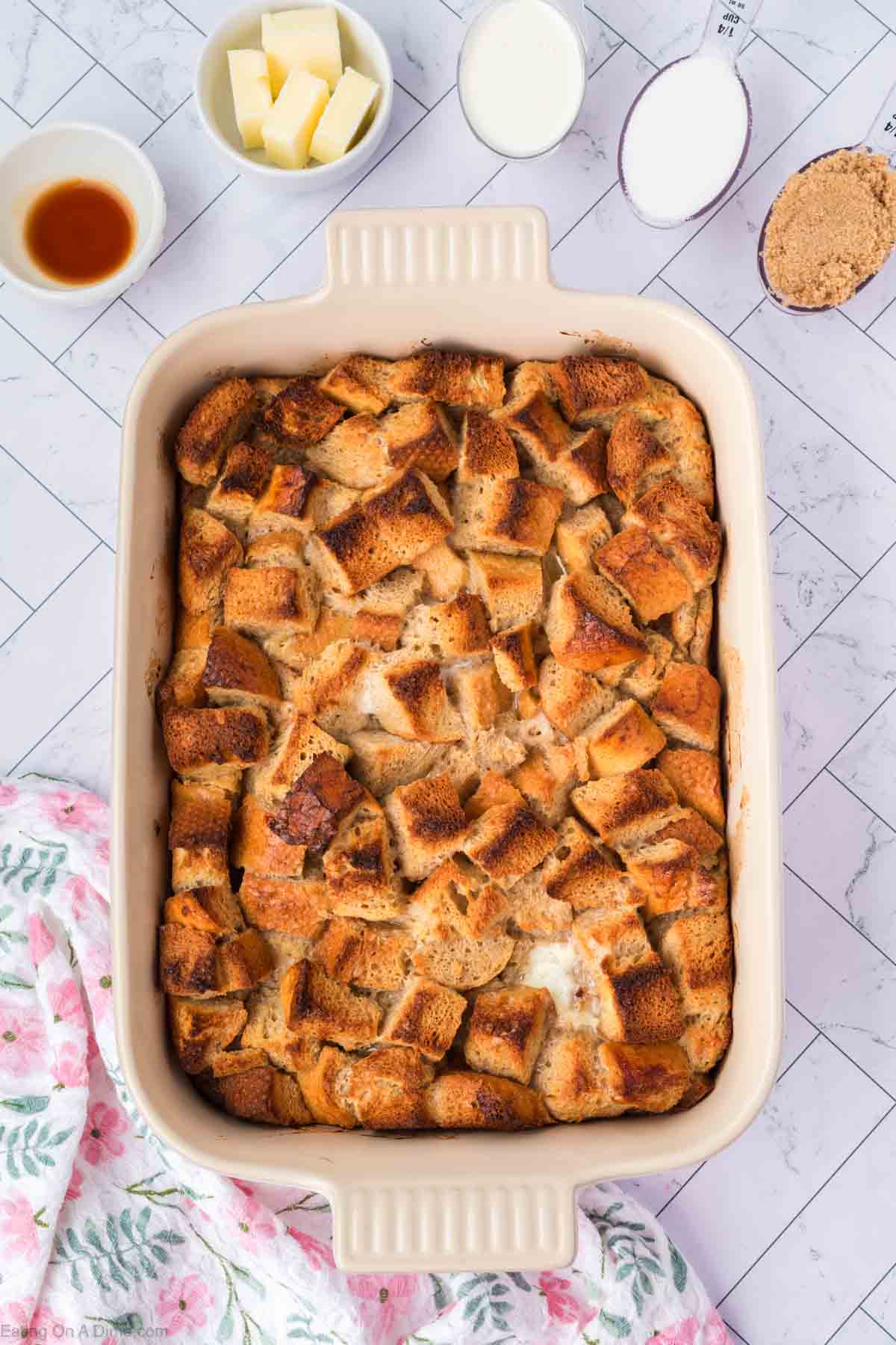 A casserole dish filled with golden-brown baked bread cubes, reminiscent of classic bread pudding, sits on a patterned surface. Surrounding the dish are small bowls with butter, milk, sugar, and brown sugar, plus a floral towel in the corner.