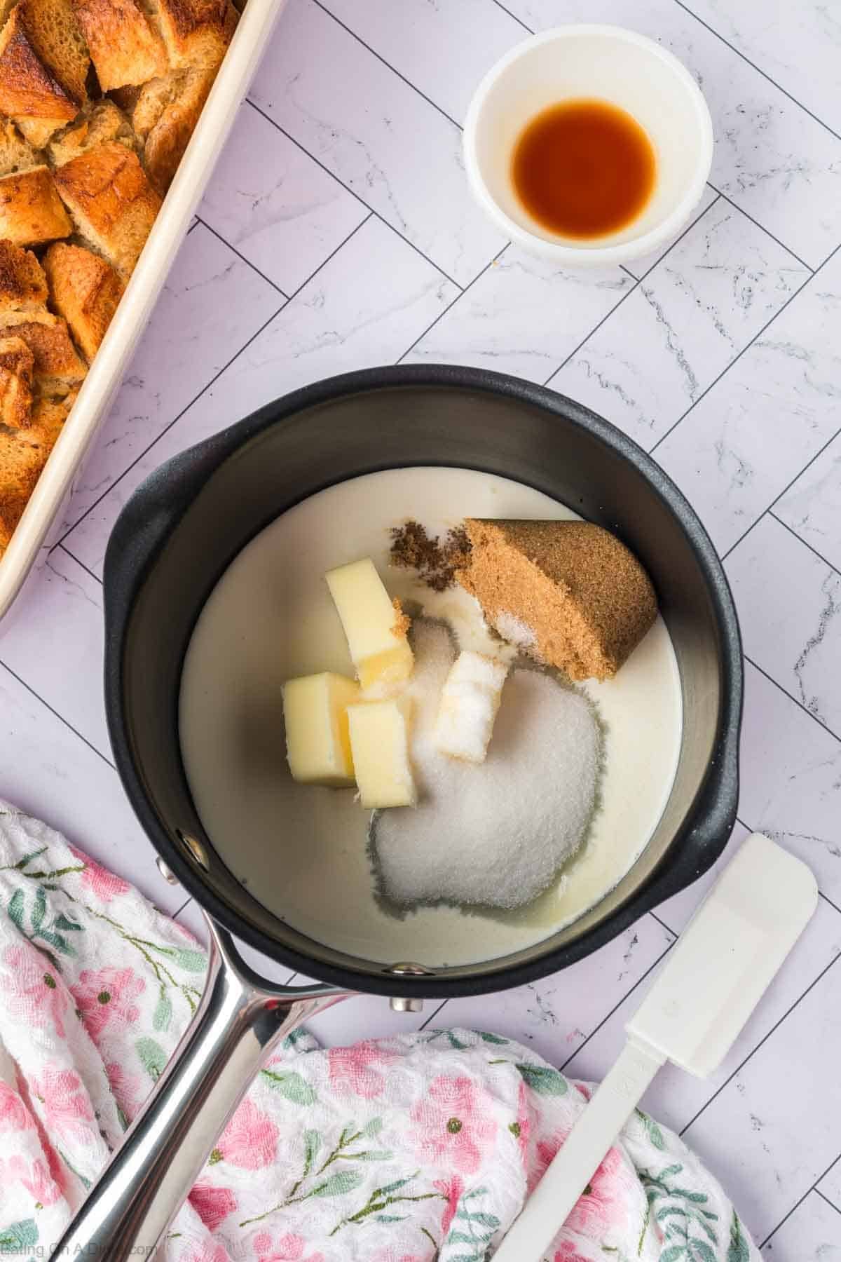 A saucepan filled with butter, brown sugar, white sugar, and milk sits on a marble countertop—ready to become a rich Bread Pudding. Nearby rest vanilla extract, a baking dish with bread cubes, and a white spatula on a floral towel.