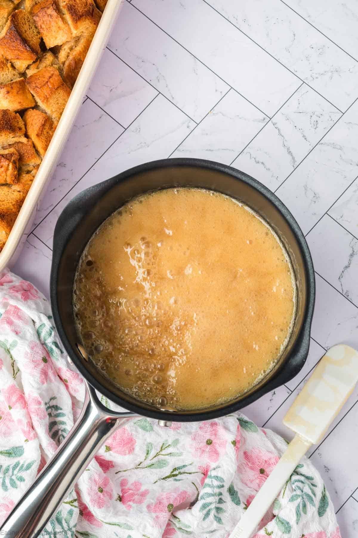 A saucepan filled with bubbling caramel sauce sits on a patterned surface next to a floral towel, with a spatula and a dish of rich bread pudding cubes nearby.