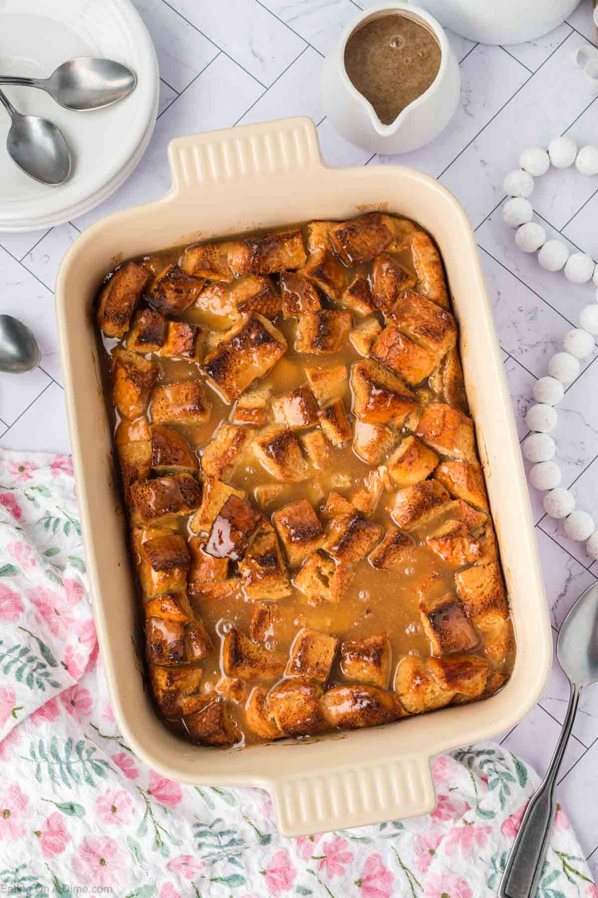 A rectangular baking dish filled with golden-brown bread pudding topped with caramel sauce, surrounded by spoons, a floral cloth, and a small pitcher of extra sauce on a white countertop—perfect for bread pudding lovers.