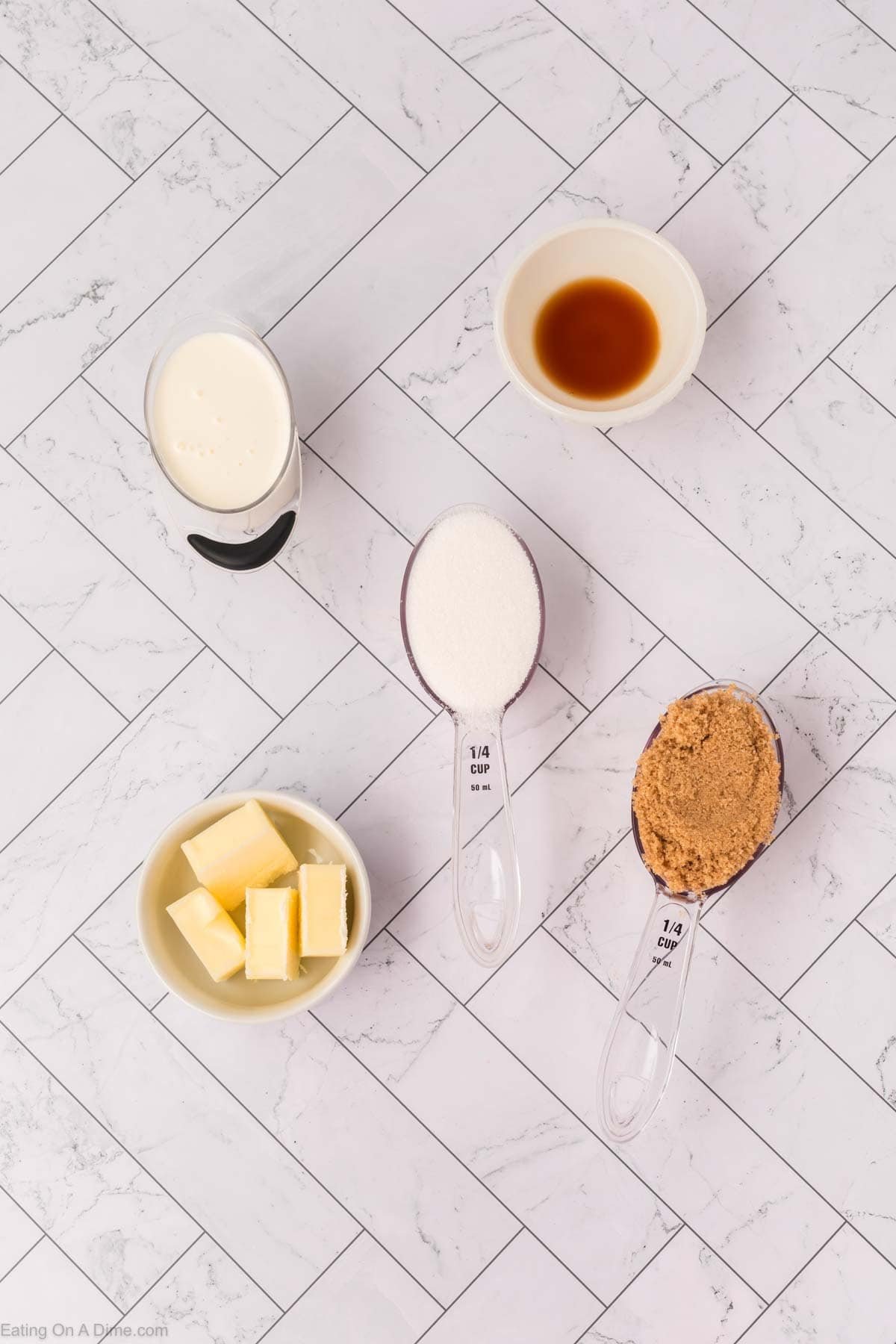 Five baking ingredients for Bread Pudding on a white tile background: heavy cream, vanilla extract, white sugar, brown sugar, and butter cubes, each displayed in separate bowls or measuring spoons.