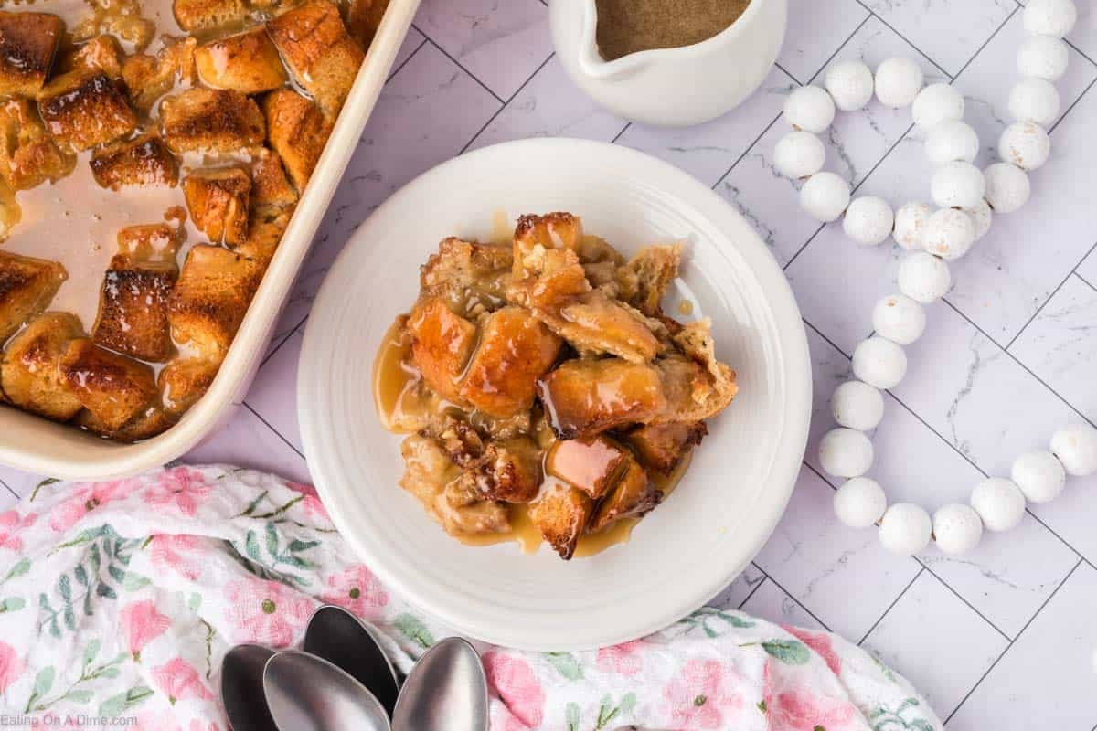 A plate of rich bread pudding topped with caramel sauce sits on a white table next to a casserole dish of bread pudding, a small pitcher of sauce, a floral towel, silver spoons, and a white beaded garland.