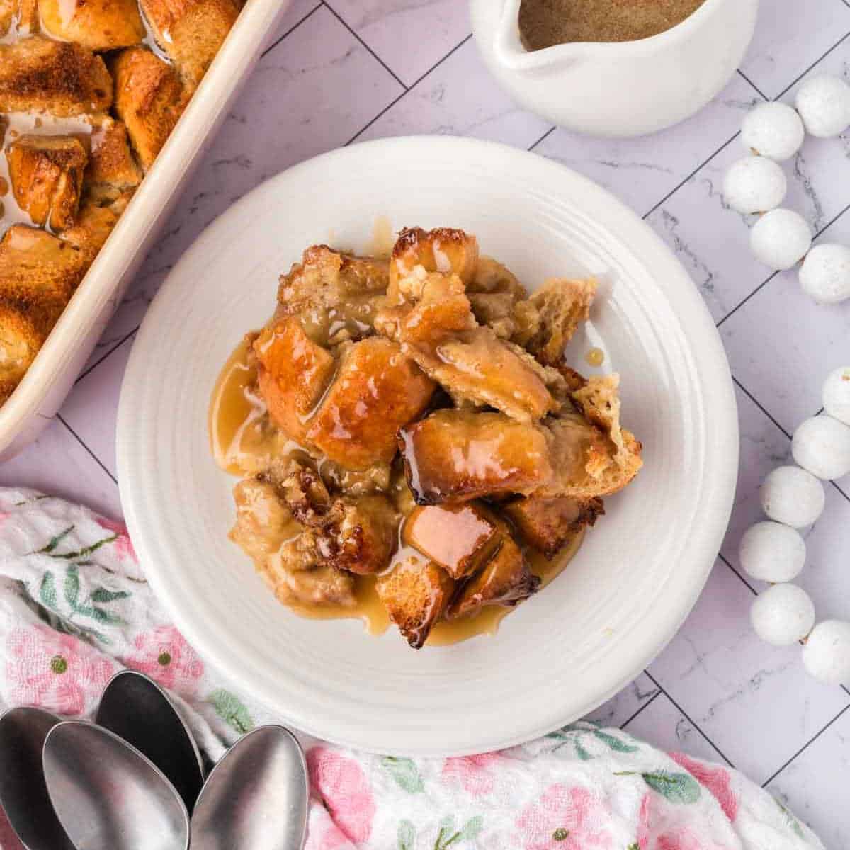 A white plate holds a serving of bread pudding drizzled with caramel sauce. Nearby are a floral napkin, three spoons, a casserole dish filled with more bread pudding, and a small white gravy boat of sauce on a marble countertop.