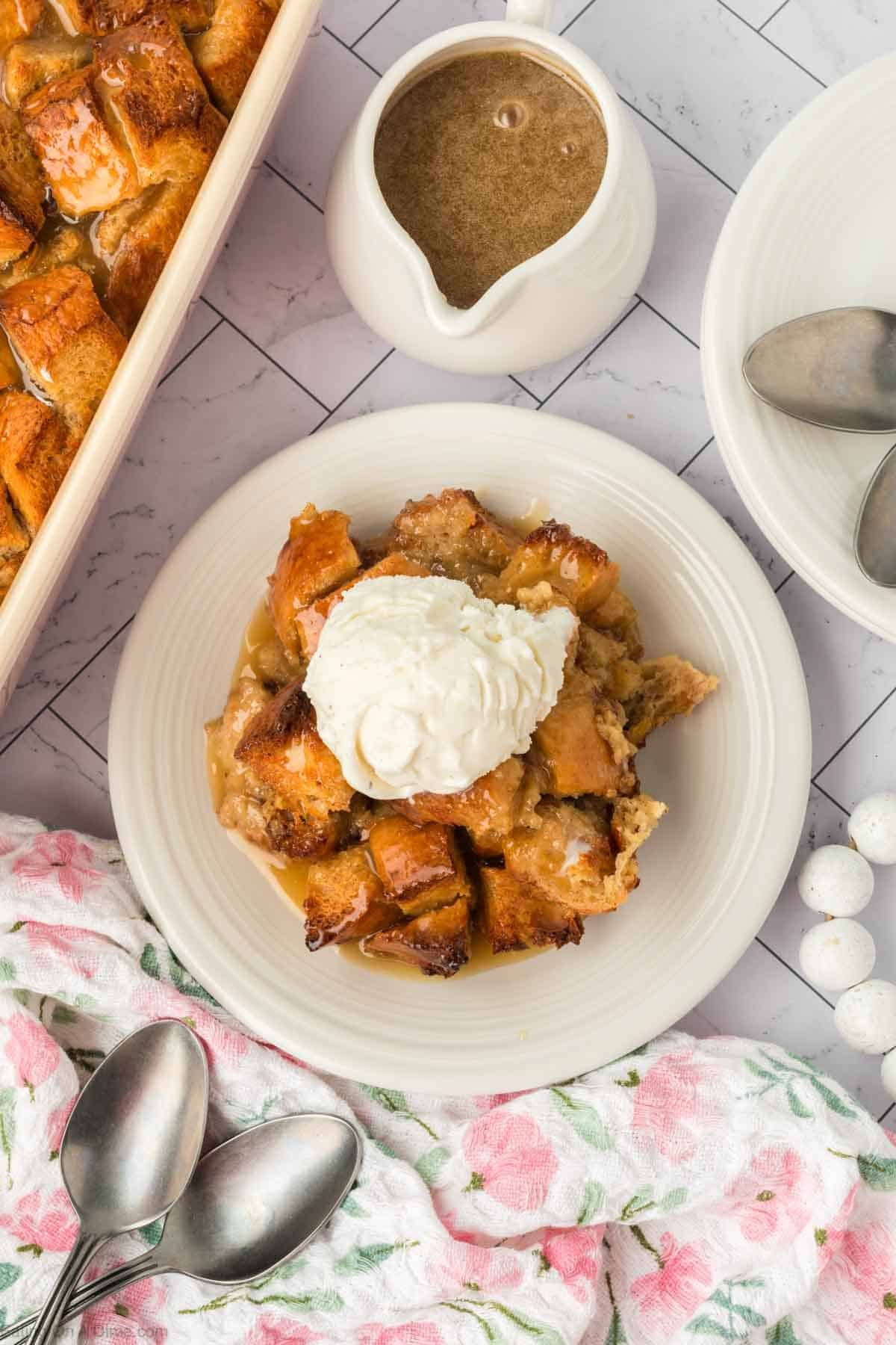 A plate of bread pudding topped with a scoop of vanilla ice cream sits next to a jug of sauce, a floral napkin, and two spoons on a tiled surface. This classic dessert is the perfect treat to share.