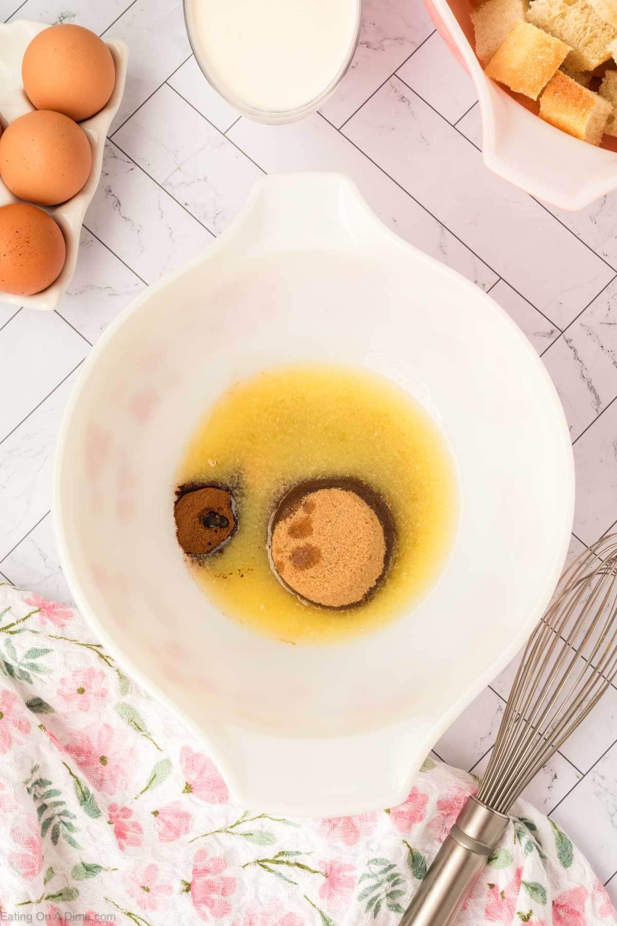 A white mixing bowl with melted butter, brown sugar, and vanilla sits on a counter near eggs, cream, a whisk, and a bowl of cubed bread—everything you need for an easy Bread Pudding dessert. A floral towel is also partially visible in the corner.