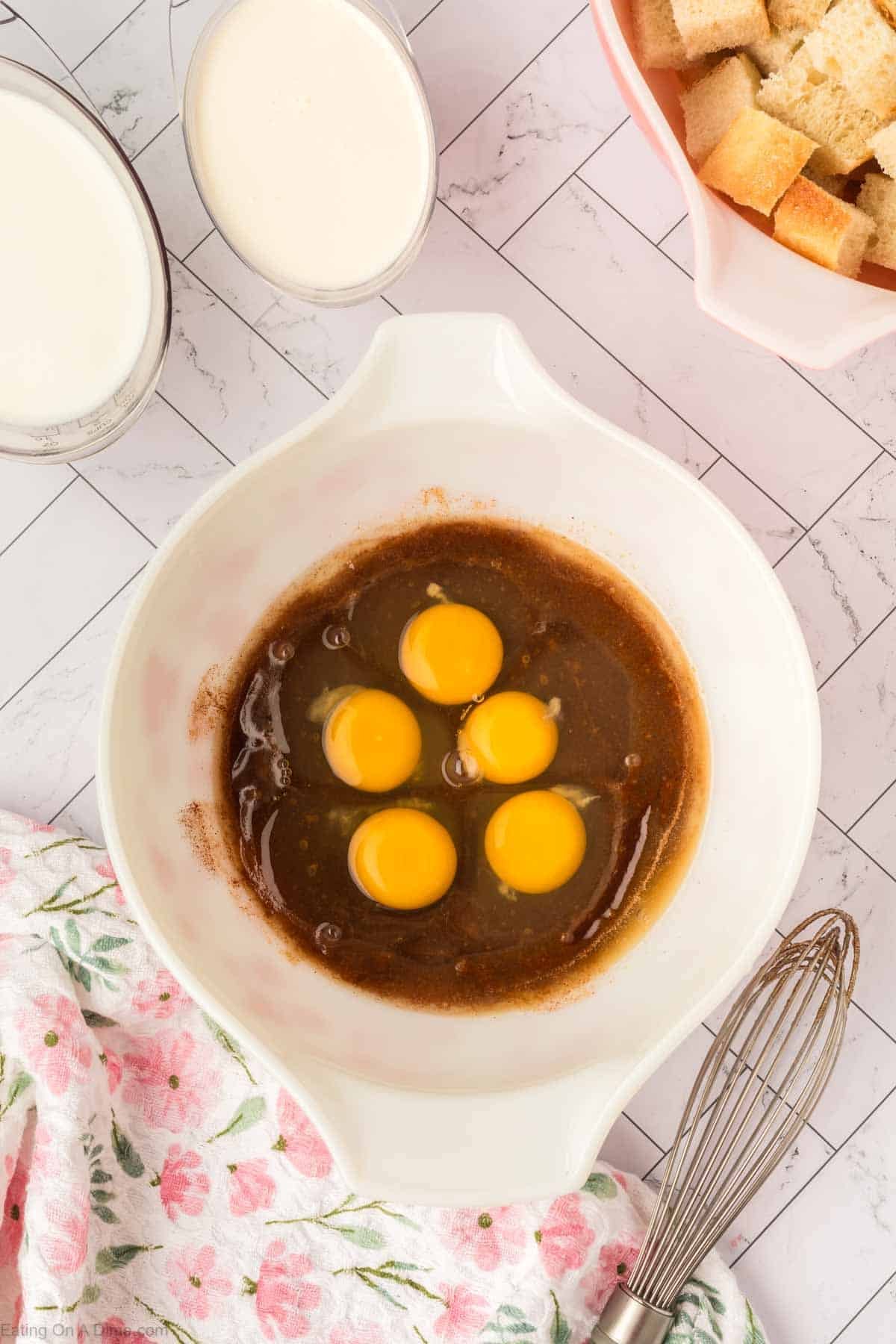 A white mixing bowl with five raw eggs and brown liquid for bread pudding sits next to a metal whisk, a pink floral towel, a bowl of bread cubes, and a glass of milk on a tiled surface.