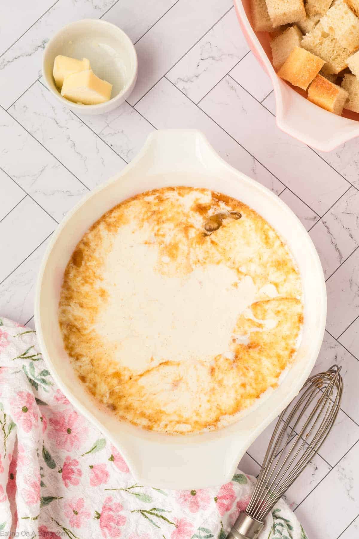 A white baking dish with creamy melted cheese, perfect for making Bread Pudding, is surrounded by a bowl of cubed bread, a small dish with butter, a floral towel, and a metal whisk on a light tiled surface.