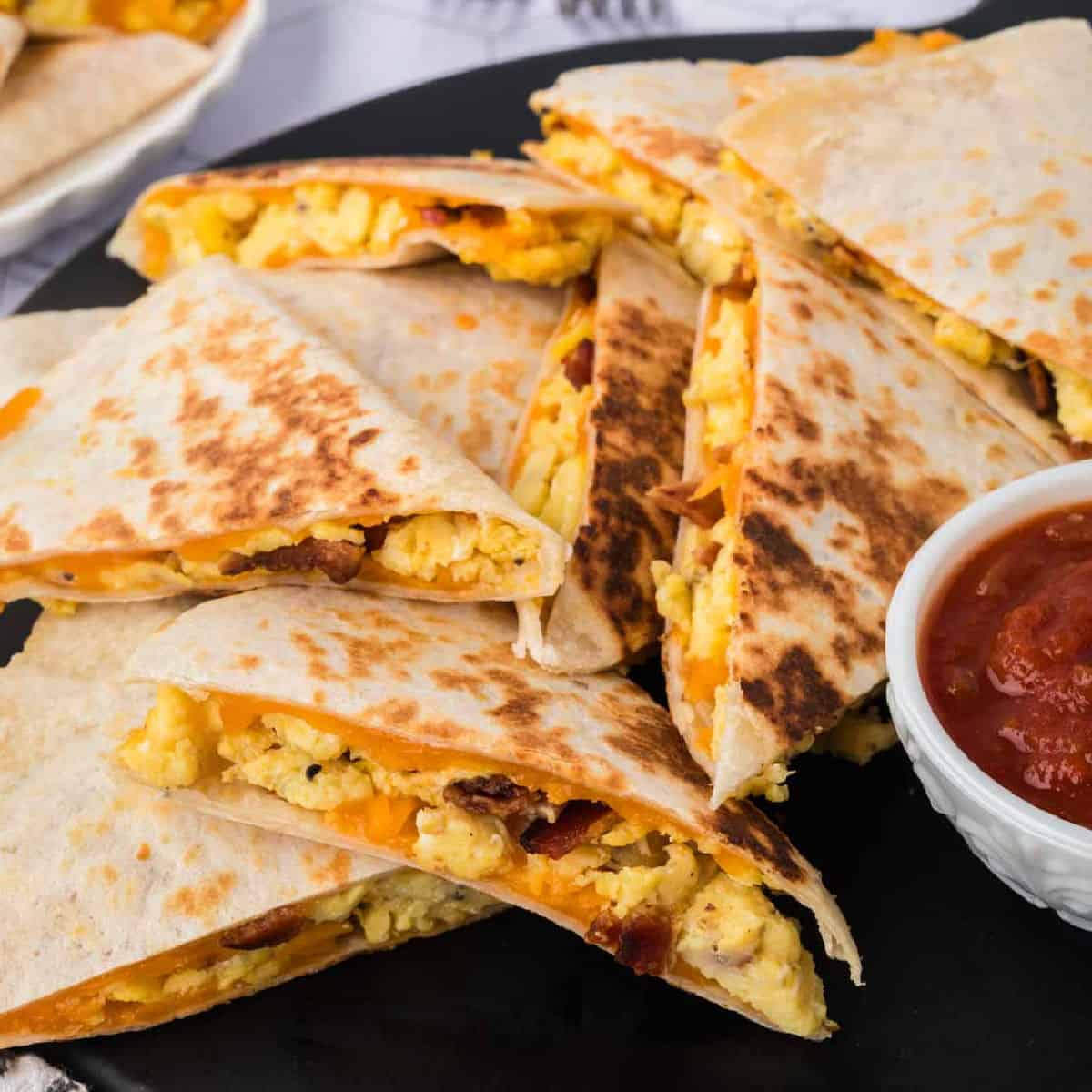 A plate of Breakfast Quesadillas filled with scrambled eggs, melted cheese, and bits of bacon, served alongside a small bowl of red salsa.