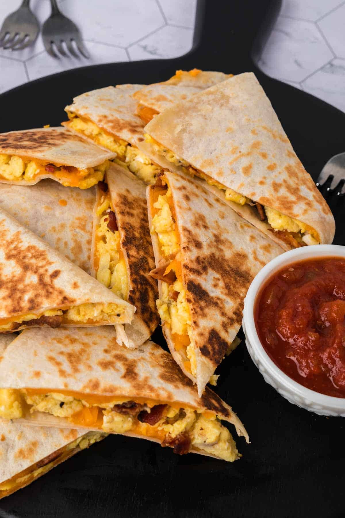 A stack of Breakfast Quesadillas filled with scrambled eggs and cheese, served on a black plate with a small bowl of salsa on the side. Two forks are visible in the background.